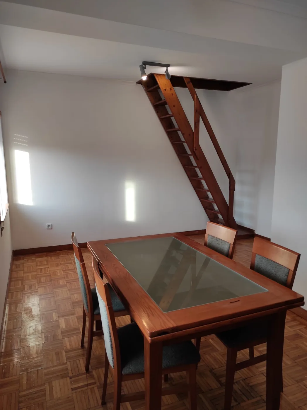 Sala do T3 com mesa de jantar de tampo em vidro e escada rebatível para o sótão, soalho em tacos de madeira